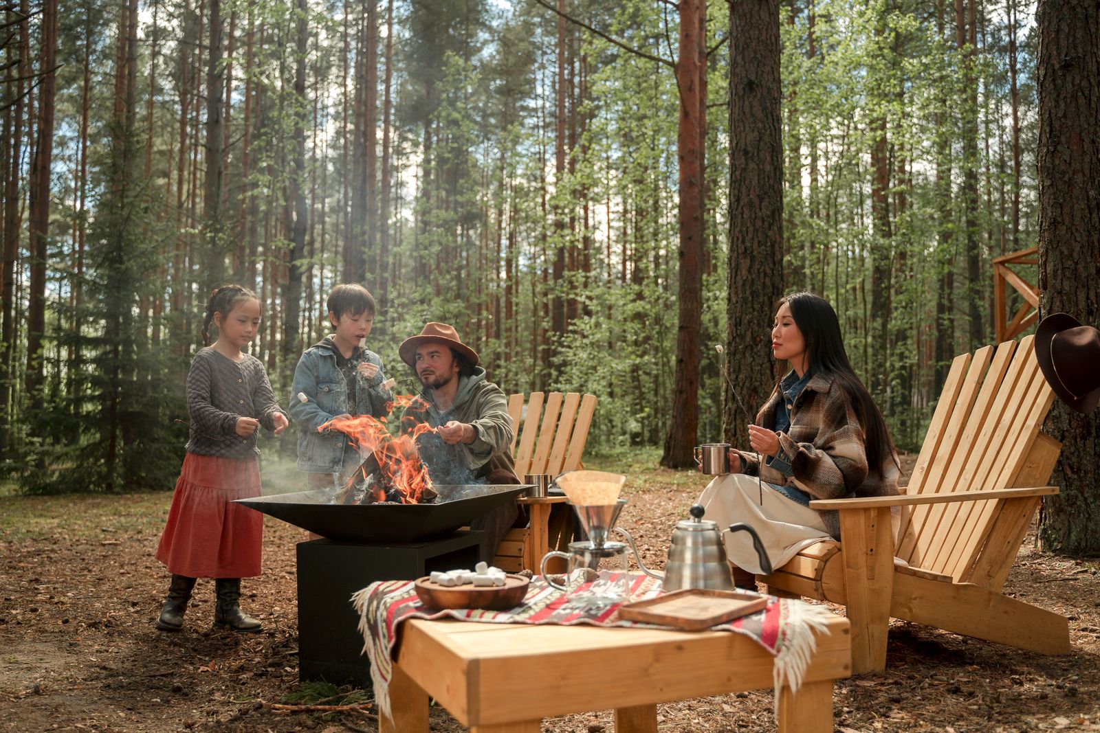 A family roasting marshmallows around a fire pit in a pine forest